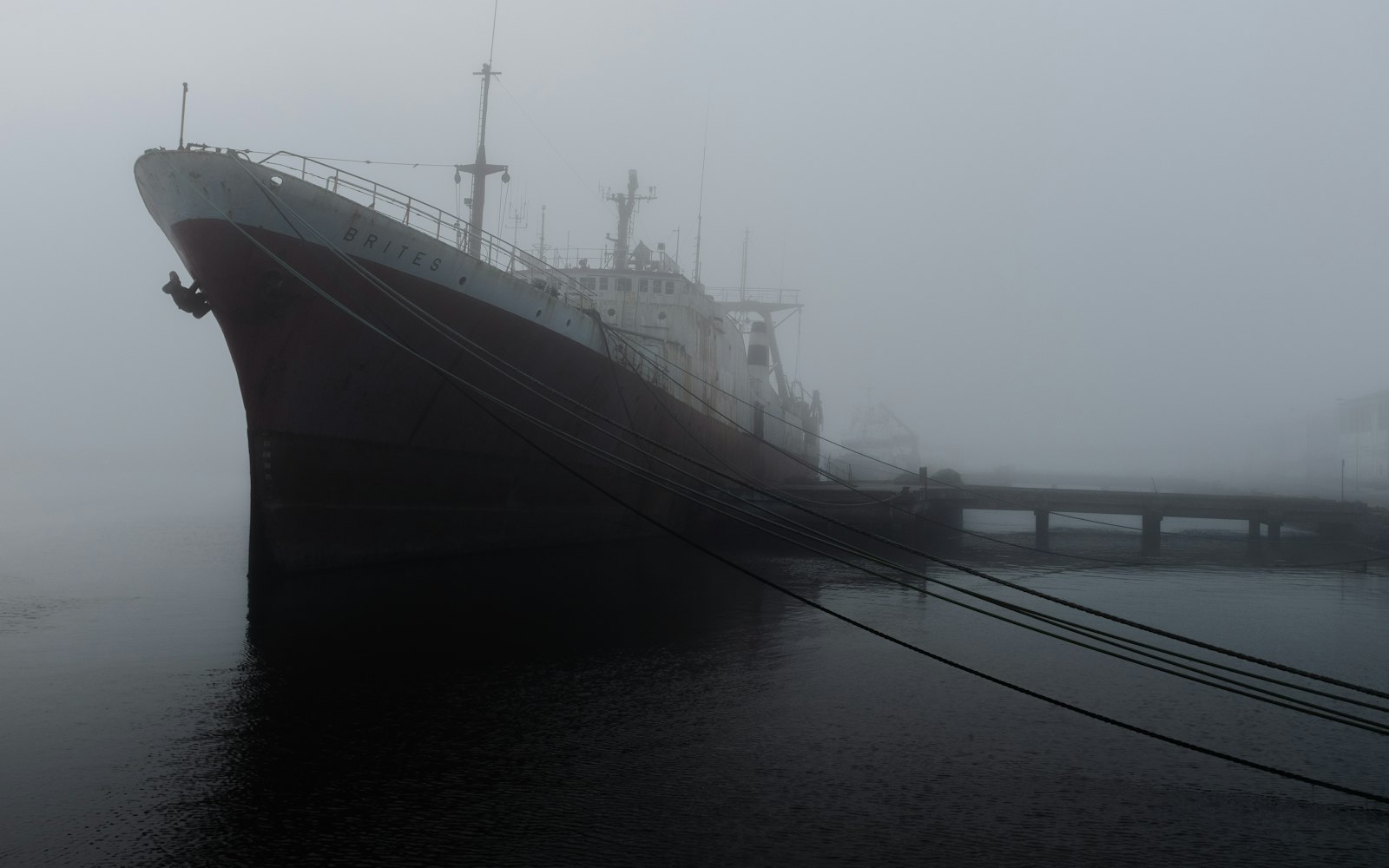 A dark red-hulled ship moored in heavy fog, its mooring lines running down to a low jetty